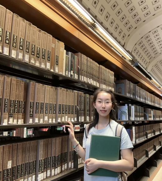A young woman standing in a library in front of a bookshelf.