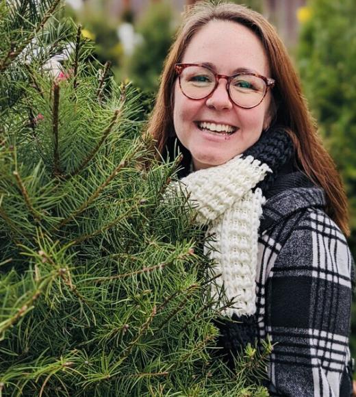 A woman wearing glasses smiling at the camera near a pine tree.