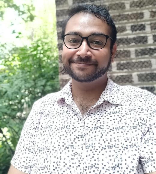 A man wearing a white shirt and glasses in front of a brick wall and greenery