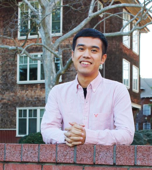 A man wearing a pink shirt this his arms folded on a brick wall