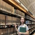 A young woman standing in a library in front of a bookshelf.