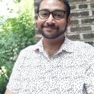 A man wearing a white shirt and glasses in front of a brick wall and greenery